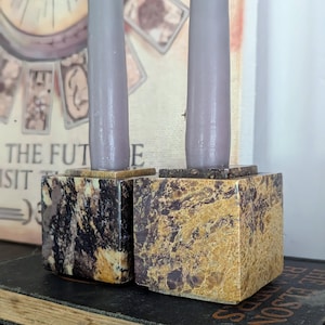 May include: Two cube-shaped candle holders made of polished stone with marbled patterns in shades of brown, black, and tan. Each holder supports a tall, smooth, light gray taper candle. The holders are positioned side by side on a dark book.