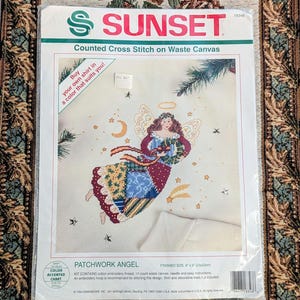 May include: A Sunset Counted Cross Stitch on Waste Canvas kit titled Patchwork Angel. The packaging features an illustration of an angel with a patchwork dress holding a wreath, with text stating Buy your own shirt in a color that suits you!