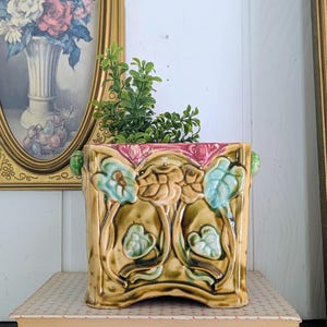 May include: A square, ceramic planter with a floral and leaf design in shades of tan, green, and blue. The planter has a pink rim and green handles. Green faux plants are inside the planter. The planter is sitting on a book.