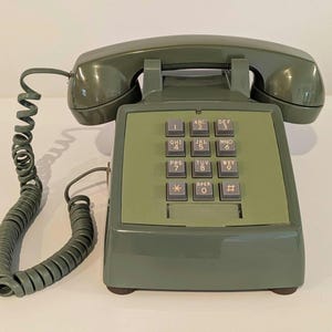 May include: Olive green vintage telephone with a handset, a coiled cord, and a keypad with number and letter buttons. The phone has a rectangular base and a curved handset cradle. The keypad includes the numbers 0-9 and special characters.