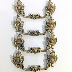 May include: Four antique brass drawer pulls with ornate, symmetrical designs. Each pull features a central, curved handle with detailed scrollwork and decorative end pieces. The pulls are arranged vertically against a white background, showcasing their aged, golden finish.