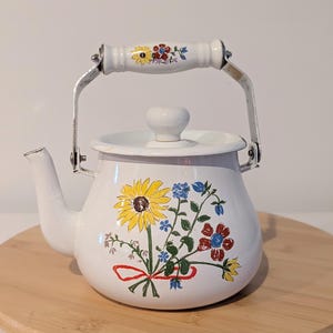 May include: A white enamel tea kettle featuring a colorful floral bouquet illustration on the front, including a large yellow sunflower and red blossoms. The kettle has a matching white lid with a knob and a white handle with floral accents.