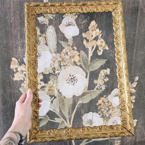 May include: A rectangular picture frame with an ornate, embossed gold finish. The frame is empty, showcasing a dark gray background featuring a printed illustration of white and muted gold flowers with green leaves and stems.
