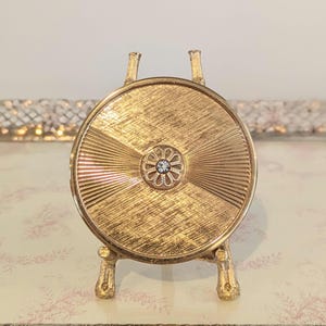 May include: A gold-toned, round compact mirror with a textured surface and a floral-shaped jewel in the center. The compact is supported by ornate legs and has two small handles. The design is vintage and elegant.