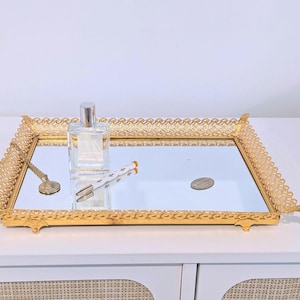 May include: A rectangular gold-toned metal vanity tray with a mirrored base and an ornate, openwork filigree border. The tray holds a clear glass perfume bottle, a white and gold pen, a gold chain necklace, and a small oval metal tag.
