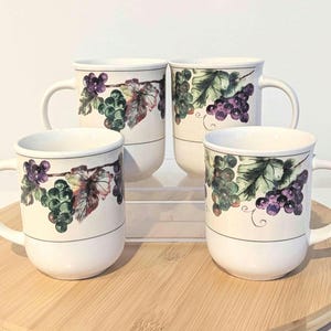 May include: Four white ceramic coffee mugs with a grape vine design. Each mug features a white base, a black stripe, and a decorative band with green, purple, and red grapes and leaves. The mugs have a curved handle.