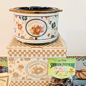May include: A white ceramic potpourri crock with a floral pattern, a dark brown lid, and a matching cardboard box. The box features the text Potpourri Crock. A clear plastic bag of Earth Harvest Simmering Potpourri Country Floral is included.