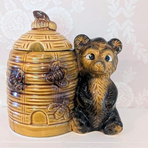 May include: A ceramic honey pot shaped like a beehive with a removable lid, featuring three molded bees on the exterior. Beside the hive sits a brown bear figurine with dark fur accents, large eyes, and a textured finish on the entire piece.