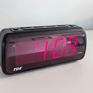 May include: A black digital alarm clock displaying the time as 4:08 PM in bright pink numerals. The clock has a rectangular shape with a dark screen and the brand name "TDE" is visible.