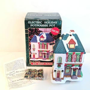 May include: An electric potpourri pot shaped like a two-story house with a dark green roof, white walls, and red accents. The packaging box is labeled ELECTRIC HOLIDAY POTPOURRI POT BAY BERRY HOUSE and includes a bag of potpourri and instructions.