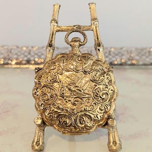 May include: A gold-colored, ornate pocket watch with a detailed, engraved design. The watch is displayed on a decorative stand with a bamboo-style design. The watch face features a detailed engraving of a ship.