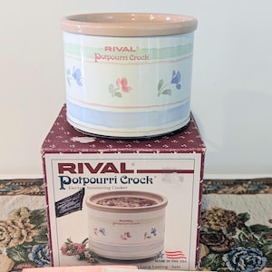 May include: A Rival Potpourri Crock electric simmering cooker with a floral pattern on a white background, featuring a light pink rim. The unit sits atop its original cardboard box, which displays the product name, image, and Made in the USA text.