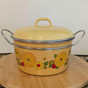 May include: A yellow enamel pot with a lid and metal handles. The pot is decorated with large yellow and red floral patterns. The pot is sitting on a wooden surface.