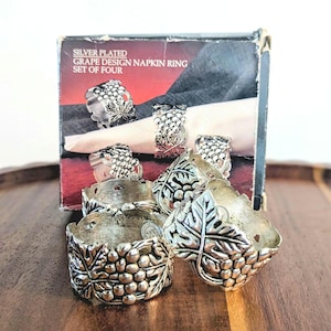 May include: A set of four silver-plated napkin rings featuring a raised grape and leaf design. The packaging box is labeled SILVER PLATED GRAPE DESIGN NAPKIN RING SET OF FOUR and displays an image of the rings holding white napkins.
