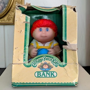 May include: A vintage Cabbage Patch Kids bank featuring a doll with red hair and blue eyes, holding a blue object. The doll is inside a worn cardboard box with the words "Cabbage Patch Kids BANK" printed on it.