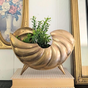 May include: A metallic gold-toned nautilus shell-shaped planter featuring a curved, ribbed exterior and three pointed legs. The vessel contains a small, vibrant green artificial plant with multiple stems and leaves, creating a contrast in color.
