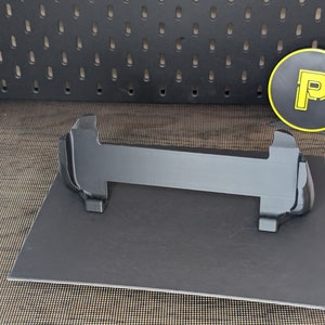 May include: A black plastic stand with two arms that curve upwards. The stand is on a black surface with a grid pattern.