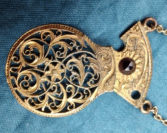 May include: A gold filigree pendant necklace with a dark red gemstone. The pendant features a delicate floral design.