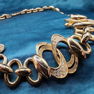 May include: A gold-tone necklace with a geometric design featuring interlocking oval and circular links. The necklace is adorned with clear rhinestones.