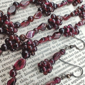 May include: A close-up of a necklace made of dark red gemstone beads. The necklace is made of two strands of beads, one strand is made of small round beads and the other strand is made of larger, faceted beads. The necklace is lying on a page of a book.