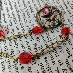 Puede incluir: Un collar de oro con un dije de ara&ntilde;a y cuentas de vidrio rojas. El dije de ara&ntilde;a tiene un strass rosa en el centro.