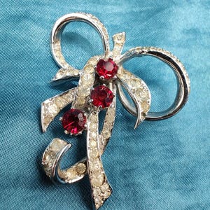 May include: A silver tone brooch with a bow design and three red rhinestones. The bow is made of clear rhinestones.