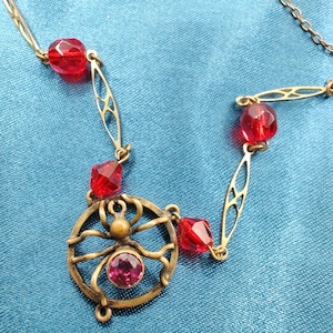 Puede incluir: Un collar de oro con un colgante de ara&ntilde;a roja. La ara&ntilde;a tiene una piedra preciosa roja en el centro. El collar est&aacute; hecho de peque&ntilde;as cuentas rojas y eslabones de cadena de oro.