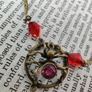 Puede incluir: Un collar de ara&ntilde;a de bronce con cuentas de vidrio rojas y rosadas. El dije de ara&ntilde;a es detallado y tiene un dise&ntilde;o de telara&ntilde;a. El collar est&aacute; sobre un libro con texto.