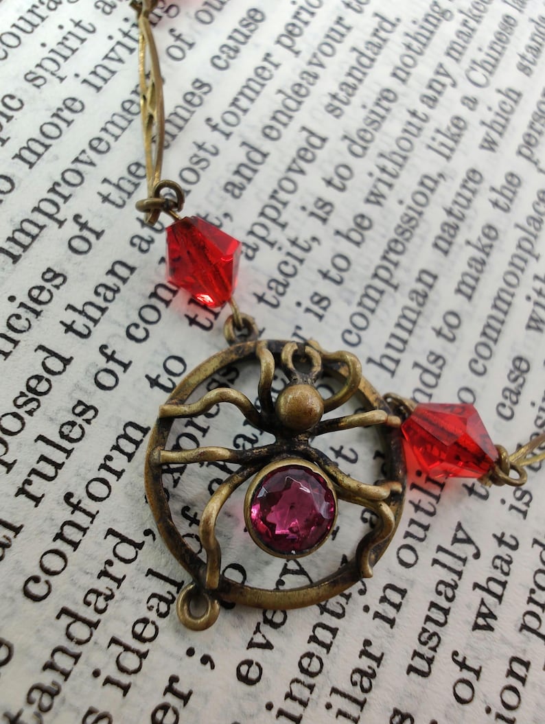 Puede incluir: Un collar de ara&ntilde;a de bronce con cuentas de vidrio rojas y rosadas. El dije de ara&ntilde;a es detallado y tiene un dise&ntilde;o de telara&ntilde;a. El collar est&aacute; sobre un libro con texto.