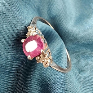 May include: A silver ring featuring a large, oval-cut, pink gemstone. The gemstone is set in a decorative setting with smaller accent stones. The ring band is plain and polished. The ring is shown on a blue fabric background.