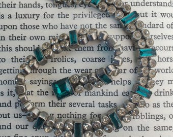Op de afbeelding: Een vintage zilverkleurige armband met afwisselend heldere en teal-kleurige rechthoekige en vierkante edelstenen. De armband is geplaatst tegen een achtergrond van gedrukte tekst.