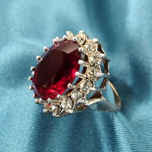 May include: A silver ring with a large, oval-cut, deep red gemstone, encircled by smaller, clear stones. The ring has an ornate setting and is set against a blue backdrop.