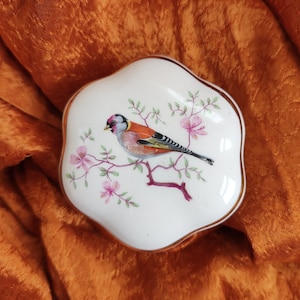 May include: A white ceramic trinket box with a gold rim and a floral design featuring a small bird with orange, black, and white feathers.