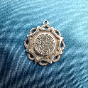 May include: A silver pendant with an intricate circular design. The pendant has a central embossed pattern, a starburst frame, and decorative openwork. A small loop is at the top for a chain.
