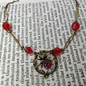 Puede incluir: Un collar de oro con un dije de ara&ntilde;a y cuentas de vidrio rojas. El collar est&aacute; sobre un libro con texto sobre filantrop&iacute;a y mejora moral.