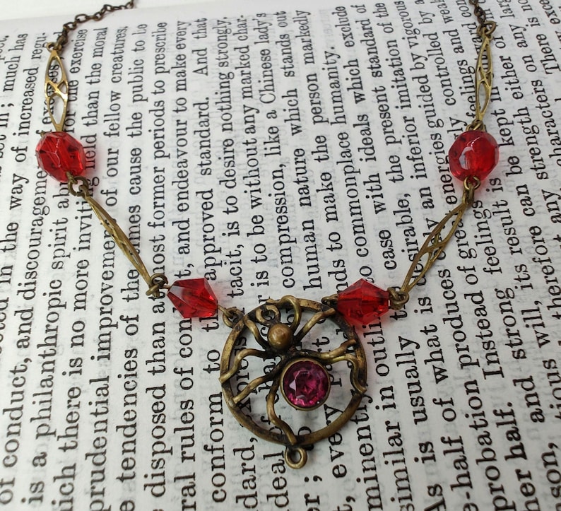 Puede incluir: Un collar de oro con un dije de ara&ntilde;a y cuentas de vidrio rojas. El collar est&aacute; sobre un libro con texto sobre filantrop&iacute;a y mejora moral.