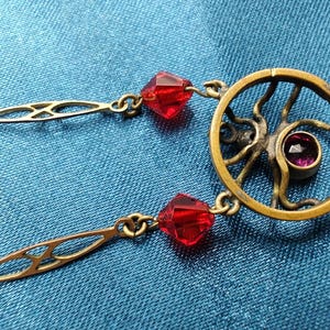 Puede incluir: Un collar de oro con un colgante circular que presenta un dise&ntilde;o estilizado y una peque&ntilde;a piedra preciosa p&uacute;rpura. El collar est&aacute; adornado con cuentas de vidrio facetadas rojas.