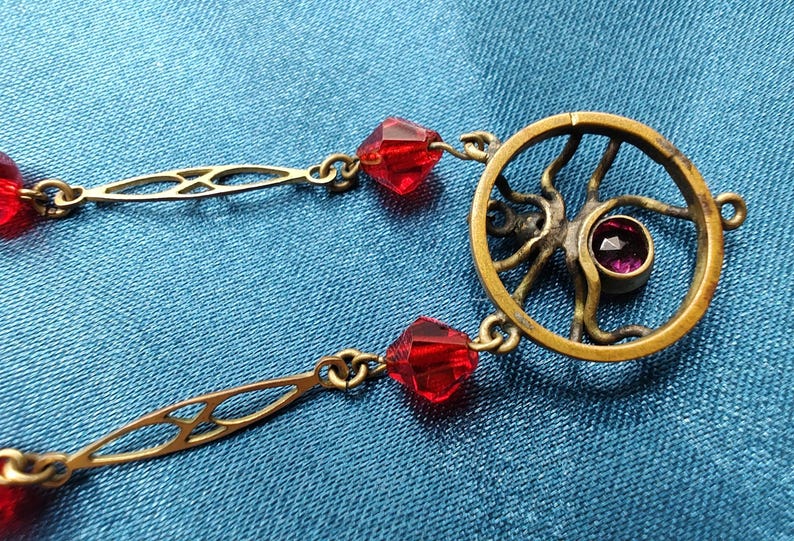 Puede incluir: Un collar de oro con un colgante circular que presenta un dise&ntilde;o estilizado y una peque&ntilde;a piedra preciosa p&uacute;rpura. El collar est&aacute; adornado con cuentas de vidrio facetadas rojas.