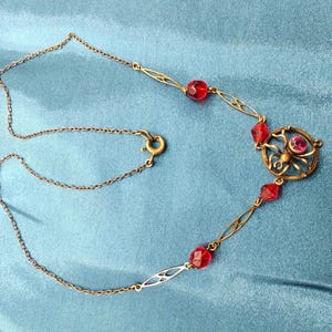 Puede incluir: Un collar de cadena dorada con un colgante de ara&ntilde;a de vidrio rojo y cuentas de vidrio rojo.