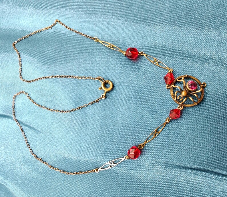Puede incluir: Un collar de cadena dorada con un colgante de ara&ntilde;a de vidrio rojo y cuentas de vidrio rojo.