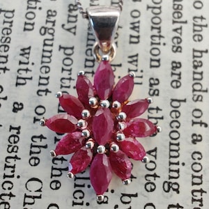 May include: A silver necklace with a pendant featuring a cluster of deep red ruby gemstones arranged in a flower-like design. The pendant is suspended from a delicate silver chain, creating an elegant and eye-catching piece of jewelry.