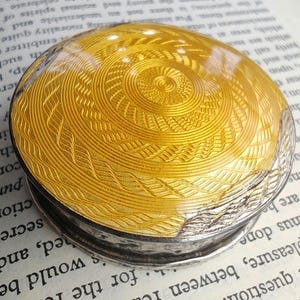 May include: A small, round, antique box with a silver base and a gold-colored, patterned lid. The lid features a detailed spiral and geometric design. The box is likely made of metal and has a reflective surface.