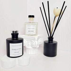 May include: Two black glass reed diffusers with labels that read "Black Pearl Scents" and the names of the scents, "Tonka & Oud Geranium" and "Mahogany Teakwood". The diffusers are on white decorative stands.