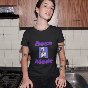Könnte beinhalten: Schwarzes T-Shirt mit den Worten "Boss Mode" in Lila und einer Kochillustration. Das Shirt wird in einer Küche mit Holzschränken und weißen Fliesen getragen.