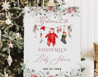 May include: A festive baby shower sign on a wooden easel, featuring a Christmas theme. The sign displays "Welcome to Samantha's Baby Shower" with baby clothes and foliage. A decorated Christmas tree and wrapped gifts are visible in the background.