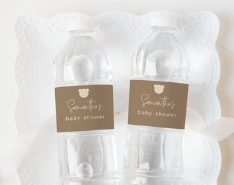 Może przedstawiać: Dwie przezroczyste plastikowe butelki z wodą z brązowymi etykietami, na których widnieje napis "Samantha's Baby Shower" i mały obrazek niedźwiedzia. Butelki znajdują się na białej koronkowej serwetce.