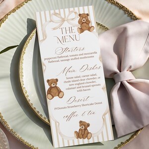 Könnte beinhalten: Eine Menükarte mit Teddybären-Motiv, mit Vorspeisen, Hauptgerichten und Desserts. Die Karte liegt auf einem hellgrünen Teller mit Goldrand, zusammen mit einer gefalteten rosa Serviette und goldenem Besteck.