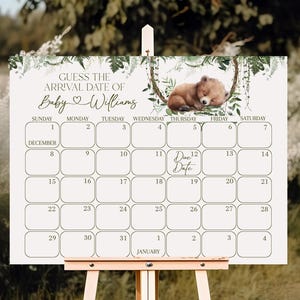 May include: A white baby shower calendar on a wooden easel. The calendar has a bear illustration and text that reads "Guess the Arrival Date of Baby Williams." The calendar grid is for December and January.