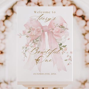 May include: A white welcome sign on a wooden easel, featuring a large pink bow with floral accents. The sign reads "Welcome to Avery's Bowtiful One" in elegant script, with the date below. The background is a blurred, light-filled room.