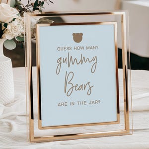 May include: A framed sign with the text "Guess How Many Gummy Bears Are In The Jar?" in elegant script. The sign has a light blue background and a gold frame. A vase of flowers and pampas grass is in the background.